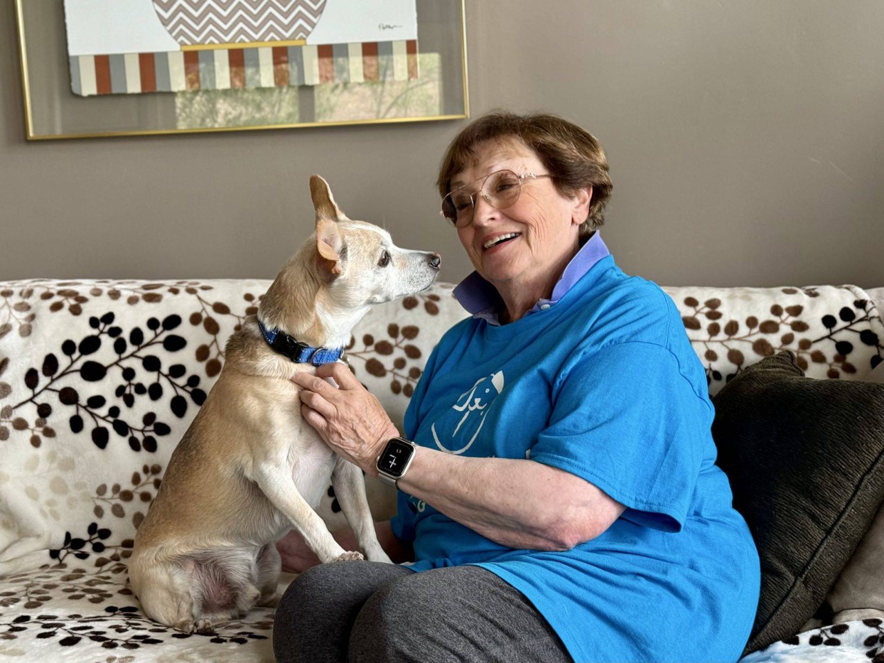 A smiling older woman in a Peace of Mind Dog Rescue shirt on her sofa with a small tan dog.