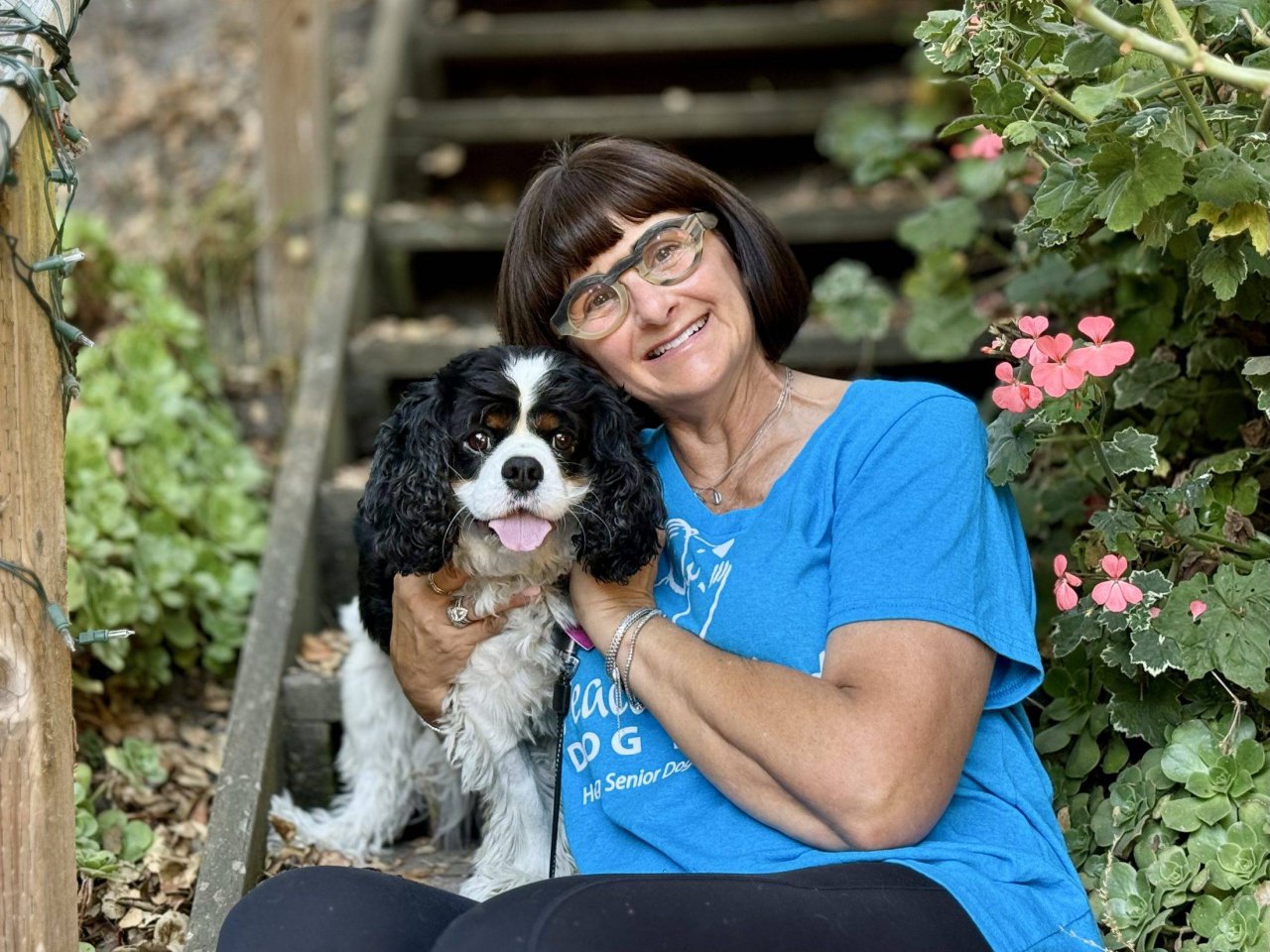 A woman in a Peace of Mind Dog Rescue shirt holding a black and white spaniel on garden steps.